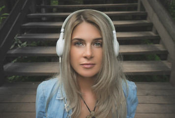 Beautiful young blond hipster woman listening music in headphones in park, sitting on wooden steps. Street fashion. A woman is wearing a denim shirt and a white summer dress