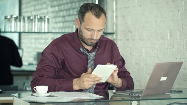 Young businessman comparing data on tablet and laptop by table in kitchen
