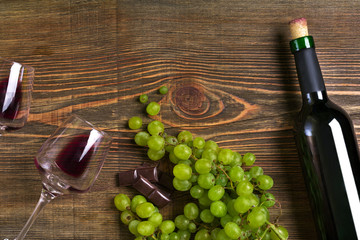 Obraz premium Red wine bottle, grape, chocolate and glasses over wooden table. Top view with copy space