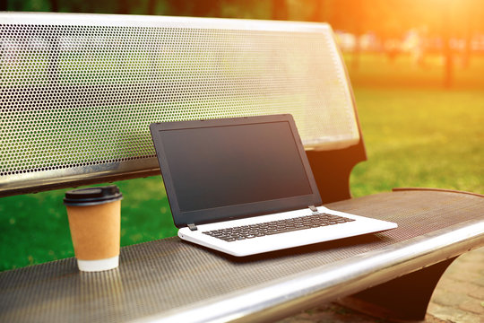 Mockup image of laptop with blank black screen and coffee cup on metal bench in nature outdoor park