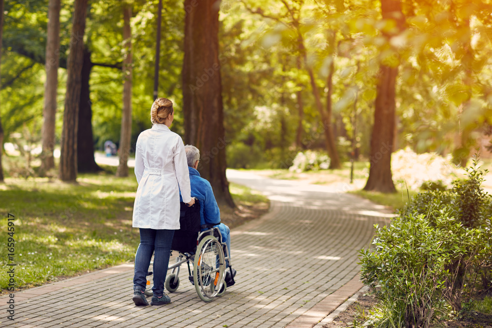 Wall mural nurse helping elderly man on wheelchair outdoor.