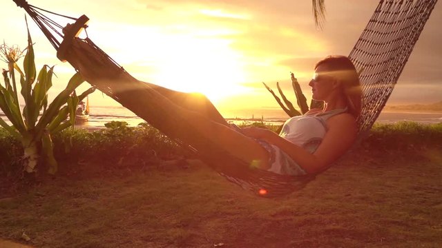 SLOW MOTION LENS FLARE: Pensive young woman on vacation laying and resting in hammock at golden sunset in hot summer. Serene woman relaxing in swinging hammock on sandy beach at sunrise golden light