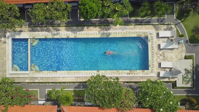 AERIAL TOP DOWN: Unrecognizable woman on summer vacation jumping head first into pool water for refreshment on hot sunny day. Girl in pink bikini diving and swimming in empty pool at luxury resort