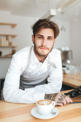 Fototapeta na wymiar Portrait of young barista in apron and white shirt dreamily looking in camera on his workplace in coffee shop. Cool boy standing at the counter with white cup of coffee in restaurant