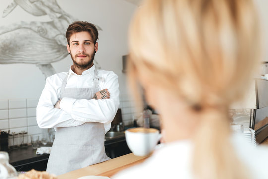 Young Barista In Apron And White Shirt Standing At The Counter With Clasped Hands And Thoughtfully Looking At Girl With Cup Of Coffe In Coffee Shop. Boy Working As A Barista In Restaurant