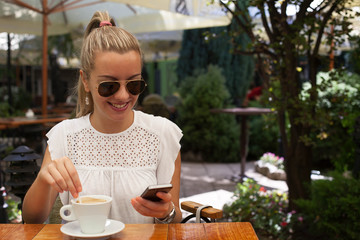 Beautiful young woman texting on the phone and drinking coffee in cafe