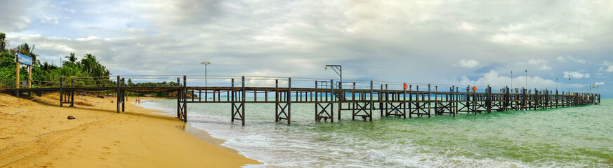 Fototapeta na wymiar Old wooden pier stretching out to sea and foamy waves on the Maenam Beach. Koh Samui information sign at entrance to pier. People are resting on the beach