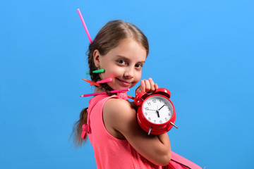 Kid in pink dress with colored pencils holds alarm clock