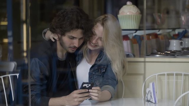 Happy young couple sitting at the restaurant looking on the screen of the smartphone and playing game on social media