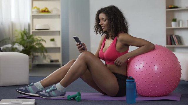 Biracial woman in gym outfit sitting on floor, using mobile phone, application