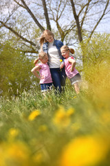 Fototapeta premium A young mother stands with her twin daughters in a flower field. The happy twins embrace their mother and enjoy the many flowers around them. 