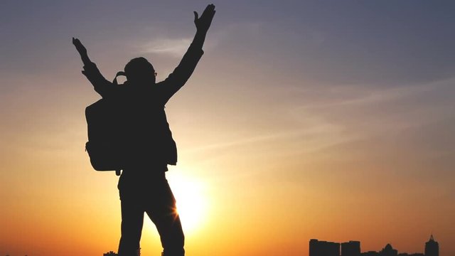 Travel Concept. Silhouette of a man with a backpack against bright sky sunset. Sun goes down. Cityscape in the background. Man looks ahead, raises his arms up. Holidays, advanture, trekking