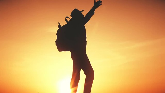 Travel Concept. Silhouette of a man with a backpack against bright sky sunset. Sun goes down. A man looks ahead, straightens his cowboy hat and raises his arms up. Orange toning filter
