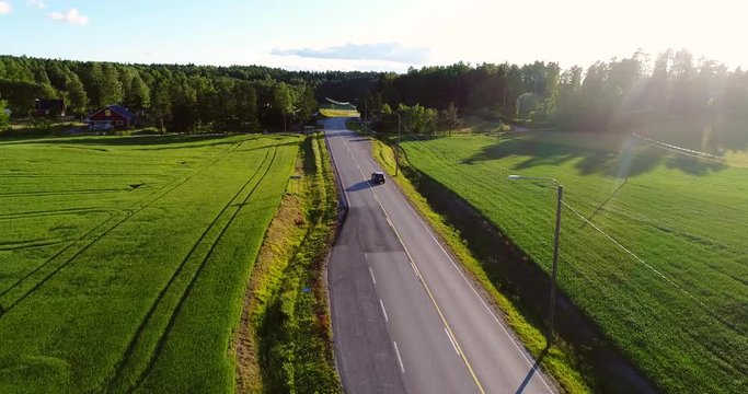 Car driving on the countryside, Cinema 4k aerial view following over a car driving on a asphalt road between yellow rapeseed fields and other green fields, at a evening, Uusimaa, Finland