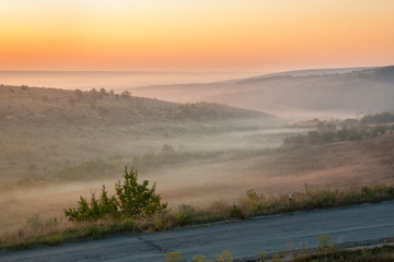 Old asphalt road in the countryside into fog at sunrise in summer.