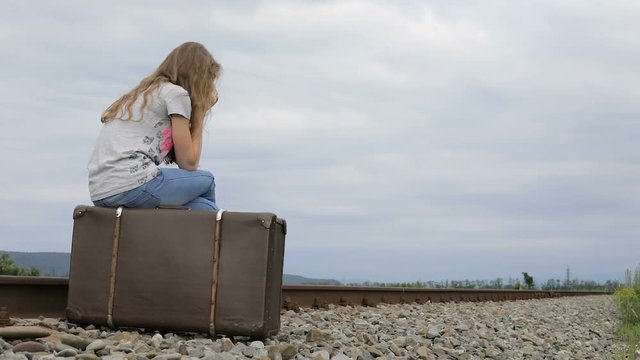 Portrait of young sad ten girl sitting with suitcase outdoors  on the railway at the day time. Concept of sorrow.