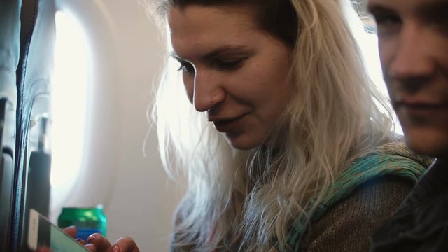 Young beautiful couple traveling by plane. Blonde woman and man sitting near the window and using smartphone.