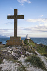 Cross in landscape of Ynys Llanddwyn Island with Twr Mawr lighthouse in background with blue sky