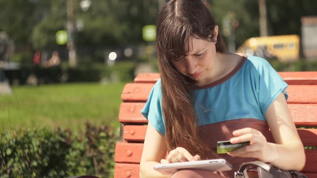 Young brunette woman using a digital tablet to pay for purchases in an online store sitting on a bench outside on a summer afternoon near the road with a busy traffic