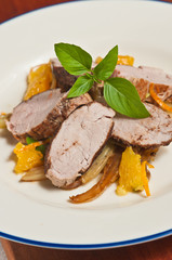 Five spices of sliced pork with orange sections and sautéed fennel with a sprig of basil on a round, white plate with a blue rim all on a wood serving board