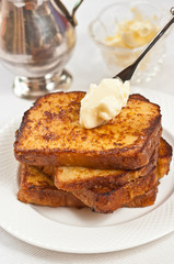 Stack of baked french toast with a artisan spreading knife with butter and warm covering the toast  and on a round, white plate  and a silver pitched  and a crystal bowl of butter