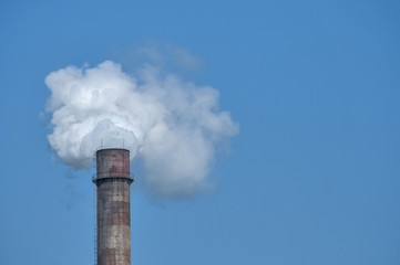 Obsolete vintage industrial chimney with white smoke on blue sky background
