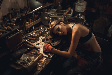 Fototapeta na wymiar Girl worker in t-shirt sits in workshop among tools and rest at her workplace.