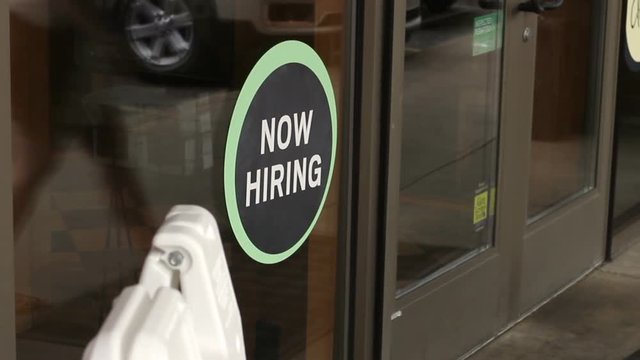 Woman walks through a door of a business with a now hiring sign in the foreground