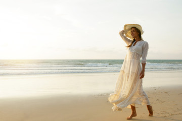 Woman wearing beautiful white dress is walking on the beach during sunset