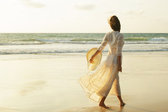 Woman wearing beautiful white dress is walking on the beach during sunset