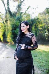 A pregnant cheerful woman stands in the park and keeps her hands on belly
