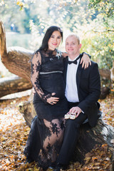 A pregnant gentle woman sits on lap of her husband who keeps a little shoes in the park