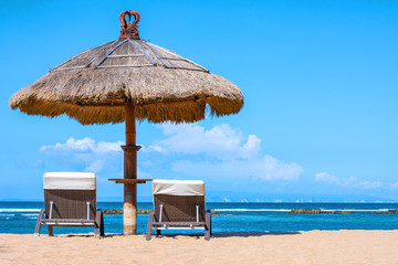 A thatched roof shady umbrella with lounge chairs on the sand overlooking a tropical beach on a prefect day.