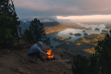 Man sitting on the rim with campfire above coastal view with islands and forests in cloudy weather from top view while sunset - 166045916