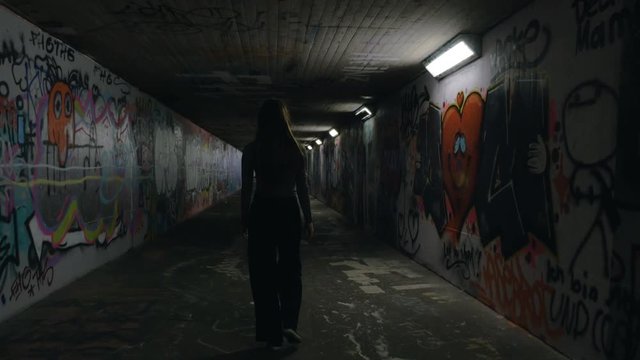 A young woman casually walks in an underground passageway with her back turned away from the camera. The underground passageway is spray painted all over, and fluorescent light illuminates the girl.