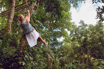 Naklejka na ściany i meble Family travel lifestyle. Happy young woman flying high with fun on rope swing on wild jungle background. Funny adventure walk in tropical rainforest. Leisure activity on summer vacation with kids.