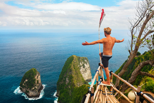 Family vacation lifestyle. Happy man with raised in air hand stand at viewpoint. Look at beautiful beach under high cliff. Travel destination in Bali. Popular place to visit on Nusa Penida island