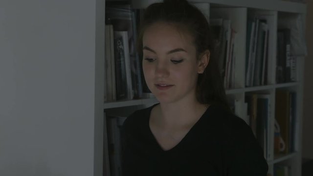 A young woman engages in reading a recital from a book. A stacked bookshelf stands behind her. Filmed from an angled profile.