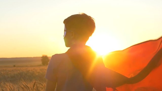 A boy with a cape of a superman stands in the golden fields of wheat looking at the horizon