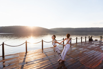 Fototapeta premium Two young women doing yoga at nature. Fitness, sport, yoga and healthy lifestyle concept - group of people making yoga pose on lake pier