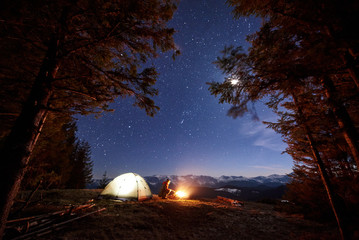 Male tourist have a rest in his camp near the forest at night. Man sitting near campfire and tent under beautiful night sky full of stars and the moon and enjoying night scene