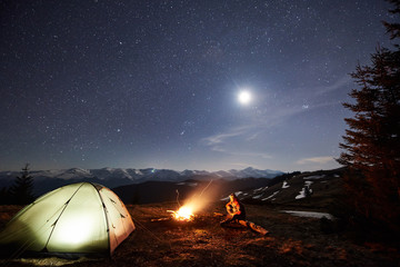 Fototapeta na wymiar Male tourist have a rest in his camp near the forest at night. Man sitting near campfire and tent under beautiful night sky full of stars and the moon, and enjoying night scene in the mountains