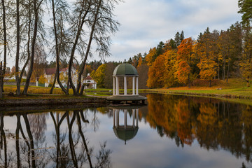 Fototapeta na wymiar Gazebo (rotunda) on the shore of pond with other resindential buildings. Estonian manor. Autumn time.