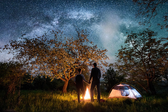Rear view of romantic couple tourists standing at a campfire, holding hands near tent under trees and beautiful night sky full of stars and milky way. Night camping