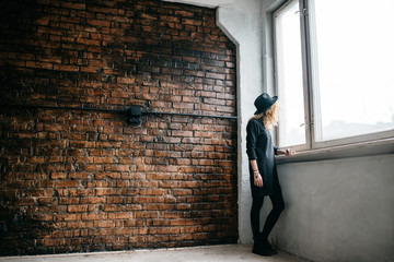 Beautiful curly girl in a bonnet standing against a brick wall And looking out the window