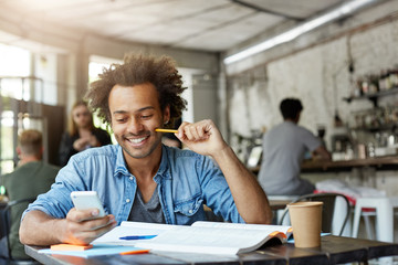Stylish Afro American male student working at cafeteria doing his home assigment having glad expression while receiving good news on his mobile phone finding out that he can be free tomorrow