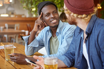 Smiling black male and his friend sitting together at pub drinking beer holding smart phones in hands communicating with pleasure. Old friends from different nationalities meeting after long time