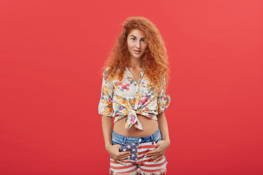 Picture of gorgeous fashionable young red-haired woman with long curly hairstyle posing in studio wearing American flag print denim shorts and cropped floral design top, showing her belly-button