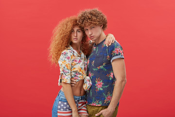 Studio shot of attractive young European couple wearing stylish clothes with floral design, embracing each other, standing isolated against red wall background with copy space for your information