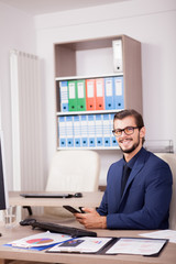 Manager with a smartphone in hands in his modern office. Image of successful businessman in blue suit. Communication and technology